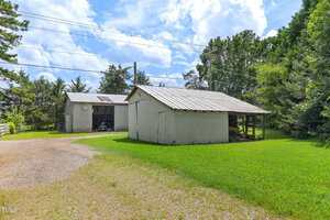 Two metal outbuildings sit on a grassy lot. One building has double doors open, revealing items inside. The other has a sloped roof and a covered area on one side. Trees and utility lines are in the background.