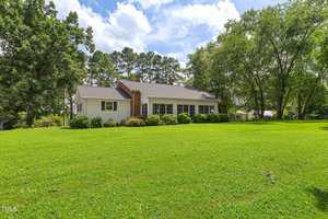 A single-story house with white siding and a brick chimney sits on a large, well-maintained lawn surrounded by trees and shrubs under a partly cloudy sky.