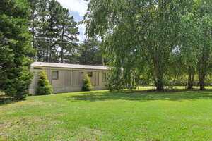 A beige mobile home sits on a grassy lawn, surrounded by mature trees and shrubs. Power lines run above the home, and the scene is lit by daylight.