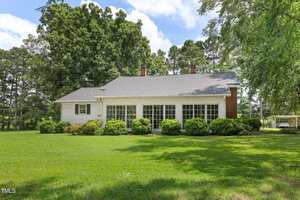 A white one-story house with a gray roof, large windows, and two chimneys sits at 2360 Warrenton Road in Henderson, surrounded by green grass, bushes, and tall trees under a partly cloudy sky.