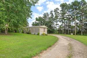 A single-wide mobile home sits on a grassy lot beside a curved dirt driveway, surrounded by trees under a partly cloudy sky.