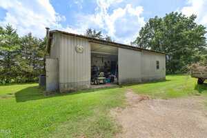 A metal shed with a partially open garage door reveals tools and equipment inside. The shed is surrounded by grass, trees, and a dirt driveway under a partly cloudy sky.