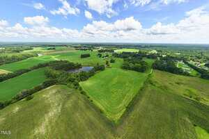 Aerial view of a rural landscape with green fields, a small pond, scattered trees, and several houses under a partly cloudy sky. Crops and open grass areas are visible, bordered by wooded sections.