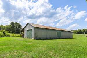 A large, rectangular metal barn with a rusted roof sits on a grassy field under a partly cloudy sky. Trees and a fence line are visible in the background.