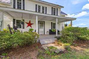 A white two-story house with black shutters and a covered front porch. The porch has plants, a bench, a rocking chair, and a large red star decor. Bushes line the walkway leading to the steps.