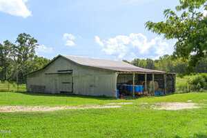 A weathered wooden barn with a metal roof stands on a grassy field. The barn has open sides with barrels, hay bales, and various equipment stored inside. Trees and blue sky with clouds are in the background.