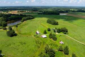 Aerial view of a rural landscape with a pond, open green fields, several barns and small buildings, a winding dirt road, and scattered trees under a partly cloudy sky.