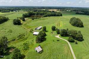 Aerial view of a rural landscape with open green fields, scattered trees, a winding dirt road, several barns and sheds, a pond, and a few small cultivated plots under a partly cloudy sky.