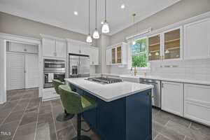Modern kitchen with white cabinets, stainless steel appliances, a large island with a built-in gas cooktop, two green barstools, pendant lights above, and a window over the sink letting in natural light.