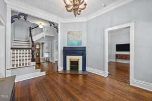 A living room with hardwood floors, a white ceiling with a decorative pattern, a chandelier, a dark blue fireplace with artwork above it, and a staircase with white spindles leading upstairs.