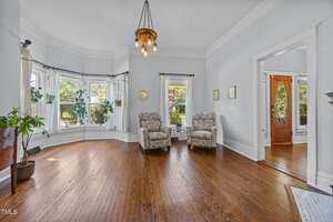 A bright living room with hardwood floors, two patterned armchairs, large bay windows with white curtains, potted plants, and a wooden entry door visible in an adjacent room.