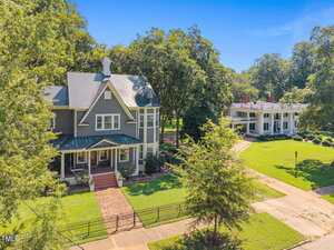 A two-story house with a covered front porch, gray siding, and white trim stands next to a wide driveway with a large white columned building in the background, surrounded by green lawns and mature trees.
