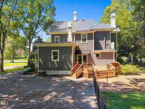 Two-story house with gray and beige siding, white trim, multiple chimneys, and a wooden deck with steps. The yard has gravel, grass, large trees, and a wire fence, with sunlight and blue sky overhead.