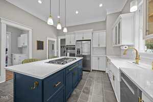 A modern kitchen with white upper cabinets, a stainless steel refrigerator, built-in ovens, a blue island with a gas cooktop, white countertops, and gray tile flooring under pendant lights.