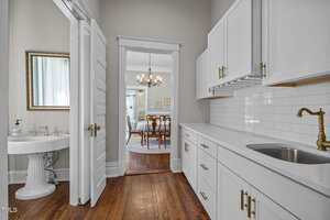 A hallway with white cabinets, brass fixtures, and a sink leads to a dining room with a chandelier. A bathroom with a pedestal sink and mirror is visible to the left. The floor is wood throughout.