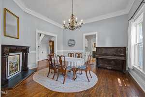 A dining room with a round table covered by a lace tablecloth, six wooden chairs, a chandelier, hardwood floors, a fireplace, a piano, and large windows letting in natural light.