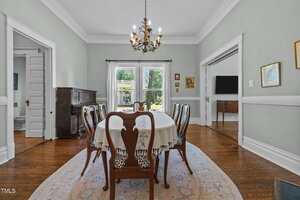A dining room with a wooden table and six chairs, a white tablecloth, a chandelier, hardwood floors, an oval rug, and large windows letting in natural light. A piano and TV are visible in adjacent rooms.