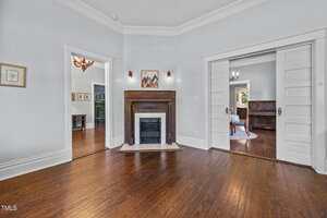 A bright room with hardwood floors, a wooden fireplace with a tile surround, white walls, and two wide doorways leading to other rooms, one with a chandelier and another with a piano visible.