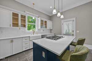 Modern kitchen with white cabinets, gold hardware, and a central island with a built-in cooktop and two green chairs. Large window above the sink, pendant lights, and neutral walls complete the space.