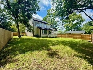 A two-story house with beige siding and white trim is shown from the backyard. The yard is grassy, fenced with wood, and shaded by trees. A small covered porch leads to the back door. The sky is partly cloudy.
