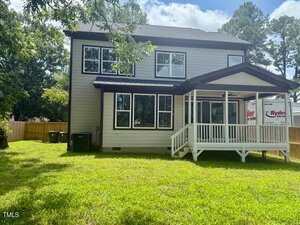 A two-story house with light gray siding, large windows, and a covered back porch. The house is surrounded by a grassy yard and enclosed by a wooden fence. A moving truck is partially visible on the right.