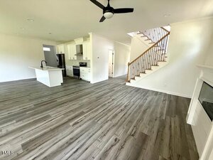 Open-concept living space with gray wood flooring, white walls, a modern kitchen with white cabinets and stainless appliances, a staircase with wooden handrail, ceiling fan, and a fireplace on the right.
