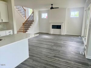 Open-concept living area with gray wood flooring, white walls, a fireplace, ceiling fan, and staircase with wooden handrail. Kitchen with white counters is partially visible on the left. Large windows let in natural light.