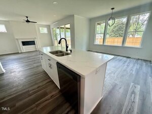A modern open-concept kitchen and living area with a white island, stainless steel sink, dishwasher, wood-look flooring, large windows, and a fireplace in the background. A dining area features a black pendant light.