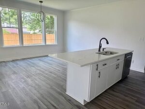 A modern kitchen with a white island that includes a sink and black faucet, adjacent to an open dining area with large windows, light-colored walls, and gray wood-style flooring.