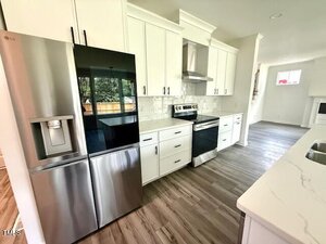 Modern kitchen with white cabinets, stainless steel refrigerator, oven, and range hood. Marble-look countertops, light-colored subway tile backsplash, and wood-look flooring. Large window reflects sunlight.