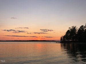 A calm lake at sunset with orange and pink hues in the sky, gentle ripples on the water, and a tree-lined shoreline on the right near 317 Doc Lane, Manson.