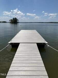 A wooden dock extends over calm lake water under a blue sky with scattered clouds. An island with trees is visible in the distance. The lower left corner displays the watermark "TMLS.