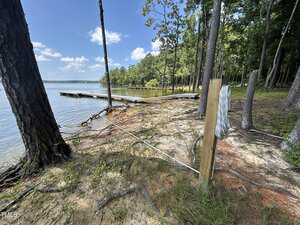 A wooden post with a white measuring tape attached stands near the edge of a forested lakeshore. Trees and grass cover the ground, and a wooden dock extends over the water under a blue sky with scattered clouds.