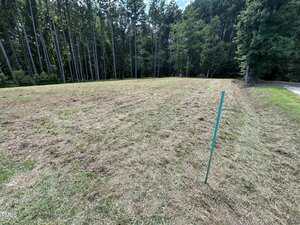 A grassy, recently mowed open field bordered by tall trees. A single metal pole is planted in the foreground, with a narrow gravel or dirt path running along the right side of the image. Sky is partly cloudy.