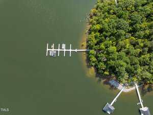 Aerial view of a dock with several boats on a greenish lake, next to a densely wooded shoreline. Another smaller dock and a boathouse are visible at the bottom right near the water's edge.