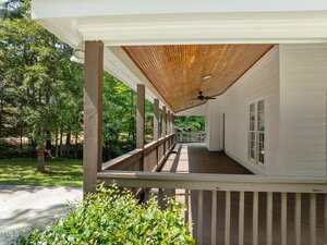A covered wooden porch with brown railings and a sloped wooden ceiling featuring a ceiling fan, attached to a white house. Trees and greenery are visible in the yard.