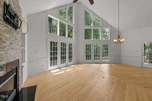 A spacious, empty living room with tall angled ceilings, large windows, and glass doors letting in natural light. The room features light wood floors, a stone fireplace, a chandelier, and views of greenery outside.