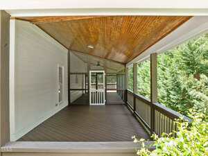 A covered porch with a wooden ceiling, dark floorboards, and light-colored walls. The porch is bordered by a railing and screened door, with trees visible outside.