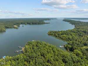 Aerial view of a large lake surrounded by dense green forest, with several docks along the shoreline under a partly cloudy sky.