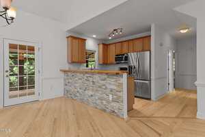 A kitchen with stainless steel appliances, wooden cabinets, and a breakfast bar with a stone facade. Light wood floors extend into the adjacent space, and a door with window panes leads outside.