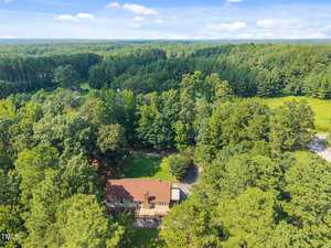 Aerial view of a two-story house with a brown roof surrounded by dense green trees, open grassy areas, and a forested landscape under a blue sky with scattered clouds.