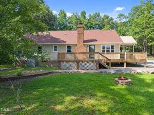 A single-story house with beige siding, a red shingled roof, a brick chimney, and a large wooden deck. The backyard features green grass, a fire pit, rocks, and trees in the background under a blue sky.