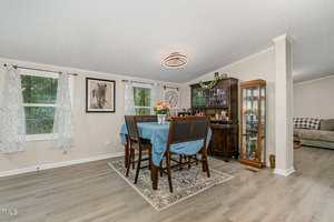 A dining area with a table covered by a blue cloth, six chairs, a floral centerpiece, a glass-front cabinet, wall art, and large windows with patterned curtains; adjacent to a living space.