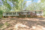 A single-story beige manufactured home with black shutters and a large wooden front deck at 4032 Royal Oak Drive, Franklinton, surrounded by trees and dappled sunlight on a dirt yard.