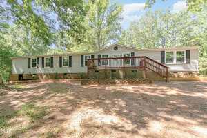 A single-story beige manufactured home with black shutters and a large wooden front deck at 4032 Royal Oak Drive, Franklinton, surrounded by trees and dappled sunlight on a dirt yard.
