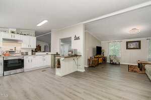 Open-plan living area with light wood flooring, white kitchen cabinets, a stainless steel stove, a breakfast bar, and a living room featuring a sofa, armchair, TV, and natural light from large windows.