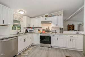 Modern kitchen with white cabinets, stainless steel appliances, a patterned rug in front of the oven, a coffee maker, microwave, and decorative items on top of the cabinets. A window above the sink lets in natural light.
