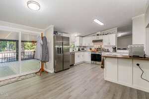 A modern kitchen with white cabinets, stainless steel appliances, and light wood flooring. A coat rack with two aprons is next to sliding glass doors that lead to a screened porch with outdoor seating.