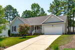 A single-story house with gray siding on 1986 Sadler Avenue, Creedmoor, features a front porch with white railing, a two-car garage, and a concrete driveway. Trees and shrubs grace the yard, with tall pine trees in the background.