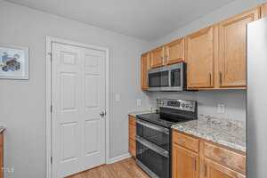 A modern kitchen with light wood cabinets, a stainless steel electric stove and microwave, marble-patterned countertops, a closed white door, and a framed picture on a light gray wall.